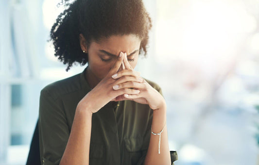 A stressed young woman with her hands pressed to her forehead, symbolizing headache, mental fatigue, and burnout in a bright office environment.