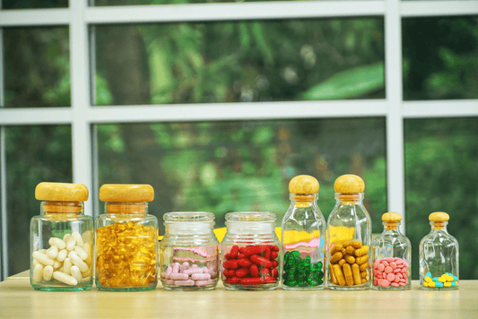 A row of small clear glass bottles containing various colors and sizes of vitamin pills and capsules, representing the problem of complex daily pill regimens.