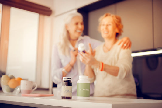 Two older women smiling while discussing vitamin bottles in a bright kitchen, suggesting bone health and joint support.