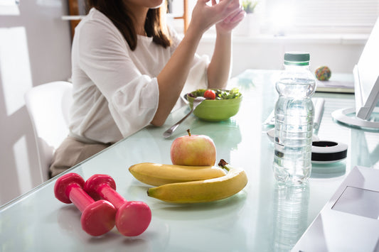 Woman managing wellness routine with healthy lunch, fruit, water, and dumbbells on a glass desk.