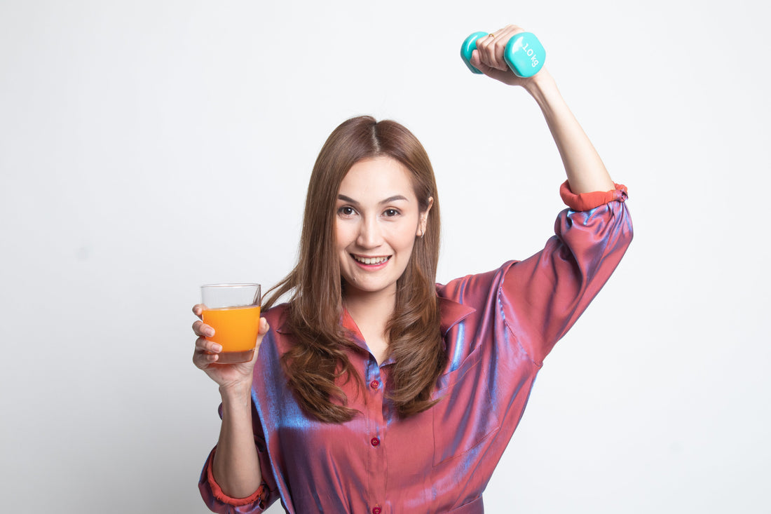 Smiling woman in a pink shirt holding a glass of orange liquid (liquid vitamins) in one hand and a dumbbell in the other, representing energy and daily fitness.