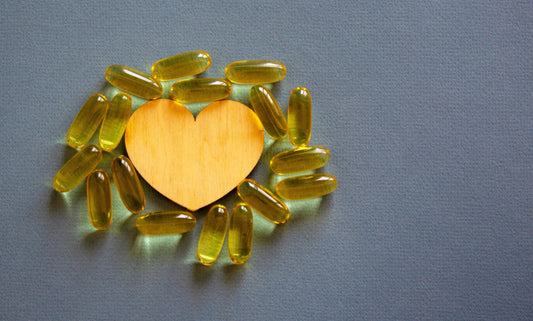 Yellow wooden heart surrounded by bright yellow Omega-3 or fish oil capsules on a gray background, symbolizing heart health and supplements.