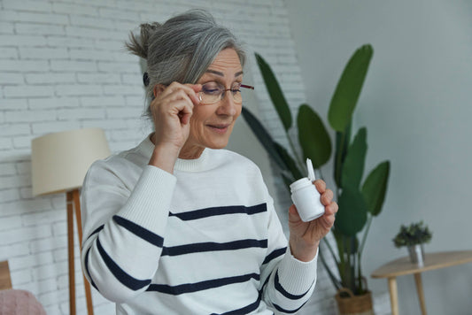 A mature woman adjusting her glasses while carefully examining a small white bottle of medicine or eye drops, suggesting focus on eye health and daily treatment.