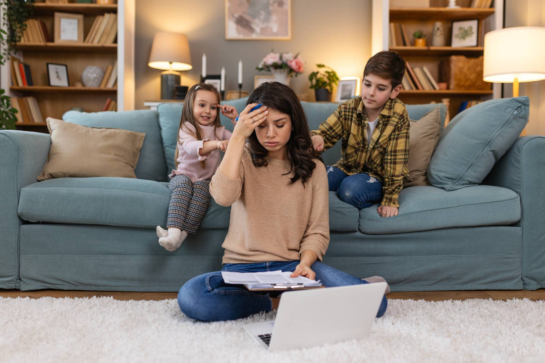 A stressed mother sitting on the floor with a laptop, holding her head while two young children demand attention on the sofa behind her, symbolizing the need for energy and focus during busy home life.