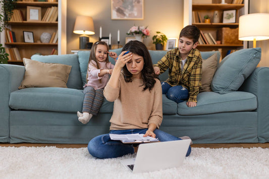 A stressed mother sitting on the floor with a laptop, holding her head while two young children demand attention on the sofa behind her, symbolizing the need for energy and focus during busy home life.