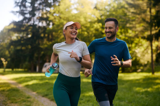 A happy, fit couple jogging on a sunlit dirt path in a park, with the woman holding a water bottle, representing active energy and outdoor wellness.