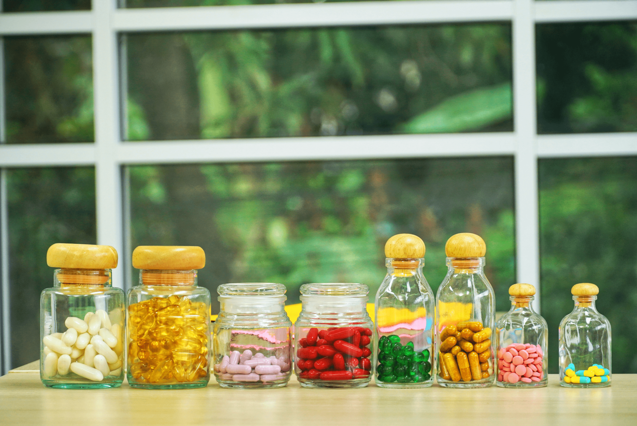A row of small clear glass bottles containing various colors and sizes of vitamin pills and capsules, representing the problem of complex daily pill regimens.