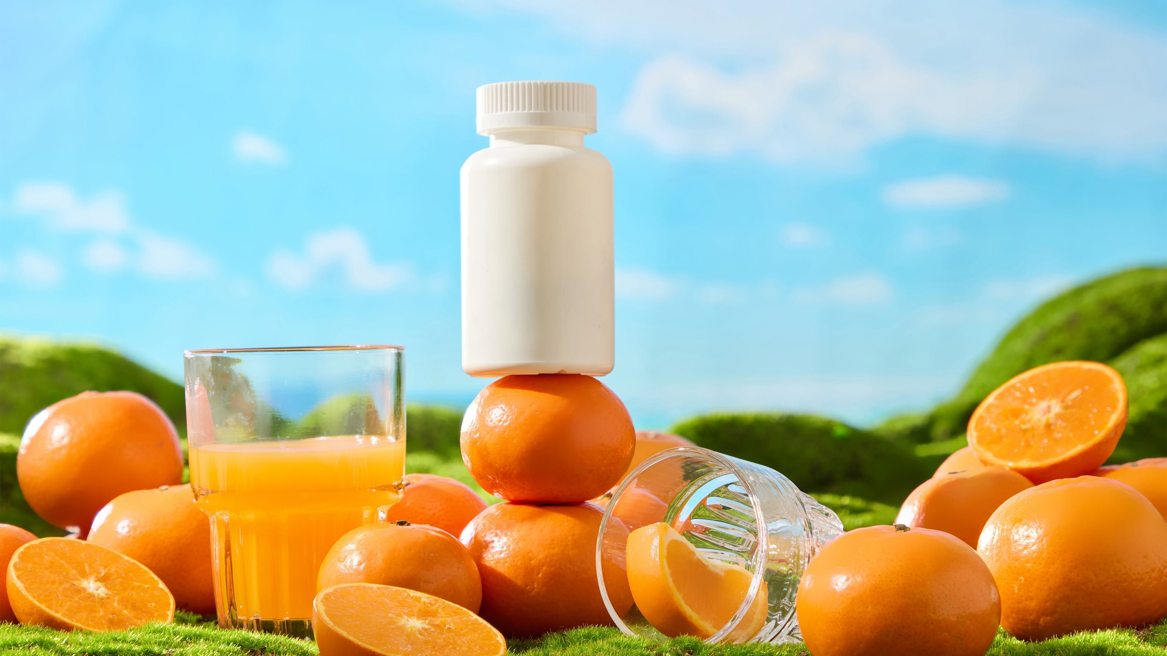 White supplement bottle balanced on a stack of fresh oranges next to a glass of orange juice on green grass, emphasizing natural flavor and liquid vitamins.