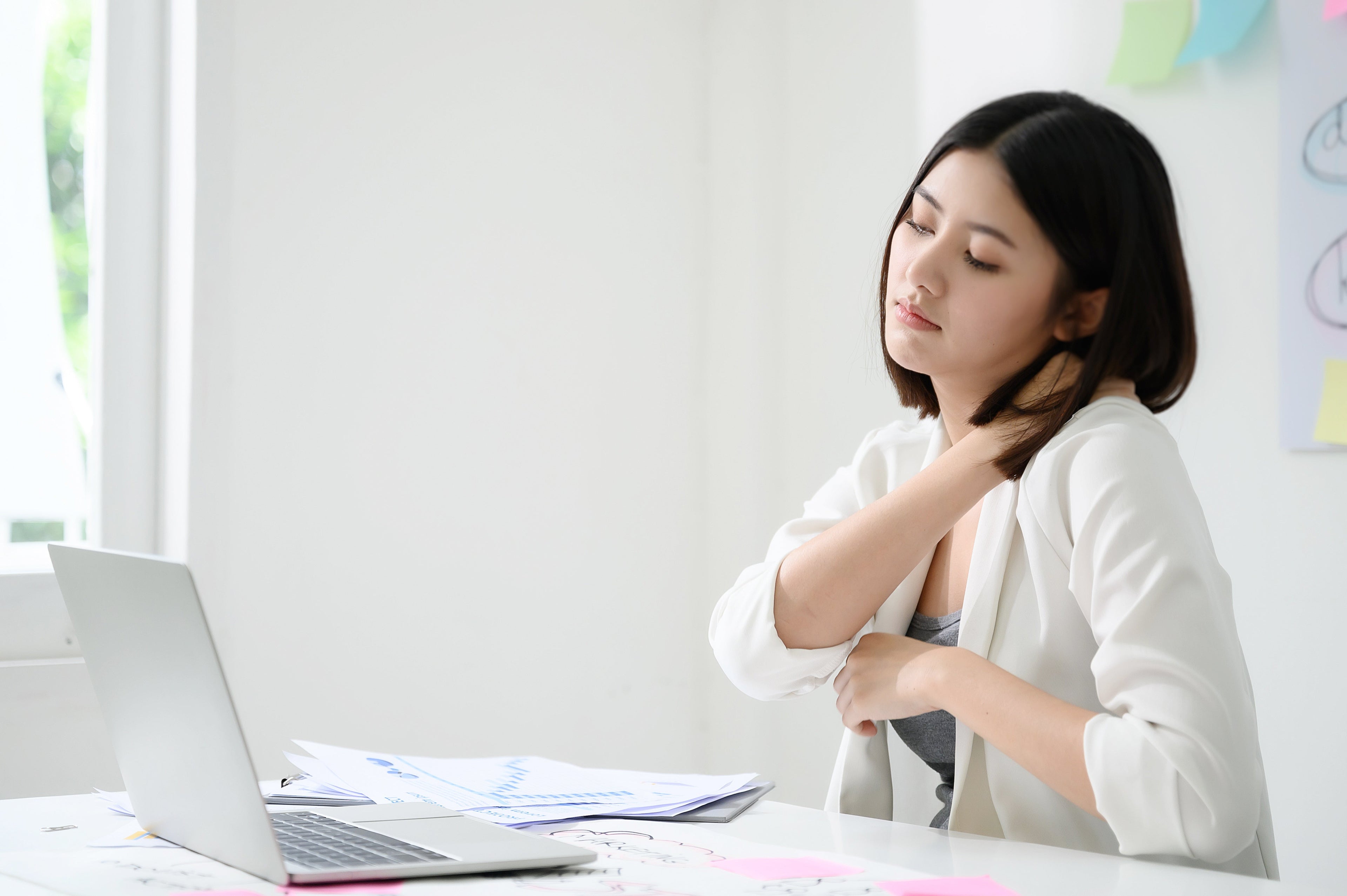 A woman in white sitting at a white table with an open laptop massaging her neck suggesting she's stressed. 