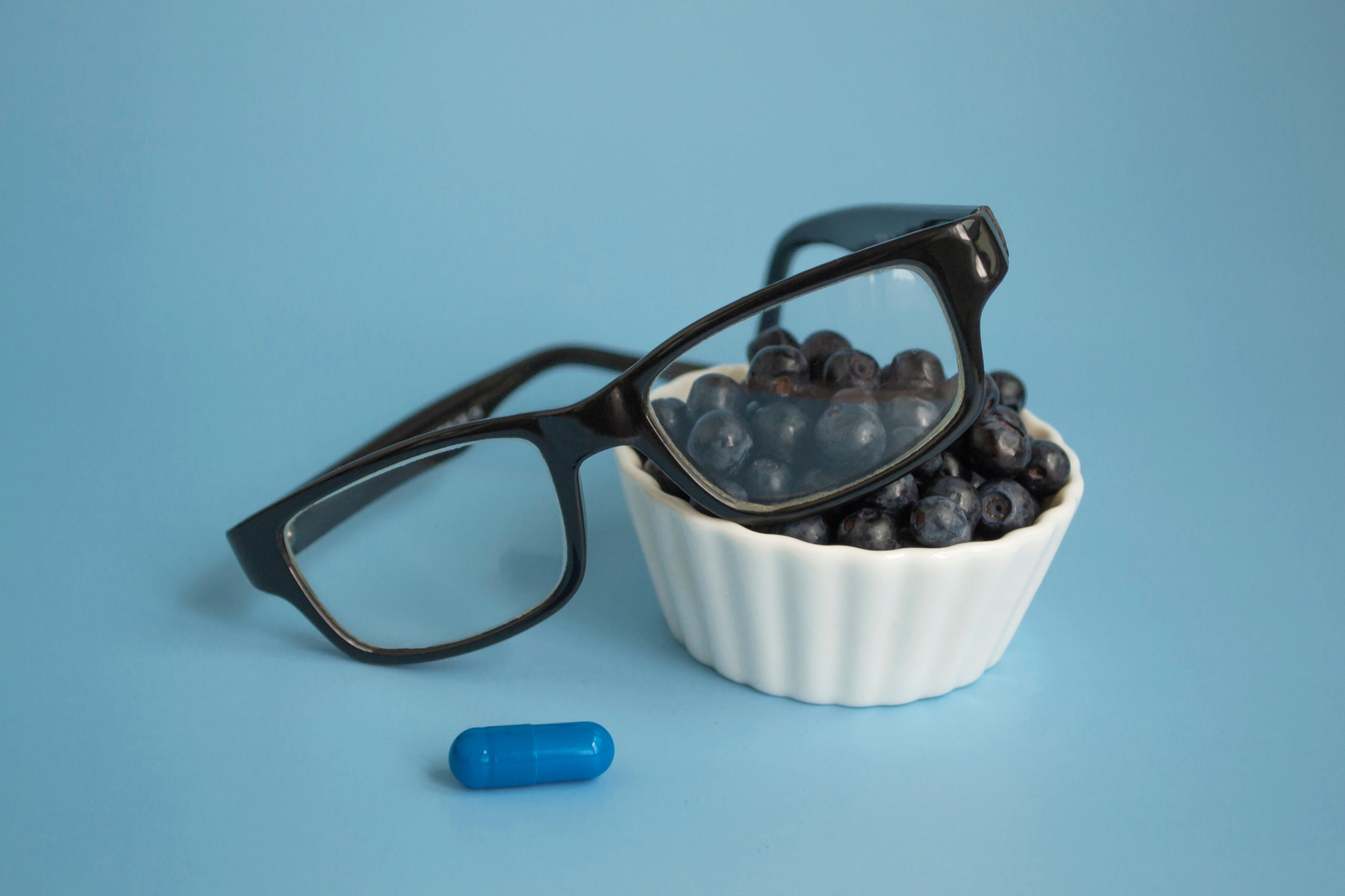Eyeglasses resting on a small white bowl of blueberries, with a single blue vitamin capsule in the foreground, representing vision health supplements and antioxidants.